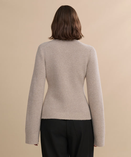 A person with shoulder-length brown hair, dressed in the Cooper Cardigan in light beige and black pants, stands with their back to the camera against a plain beige background.