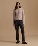 A woman stands against a warm beige studio background wearing a light taupe knit cardigan with dark buttons, black jeans, and black slip-on shoes.