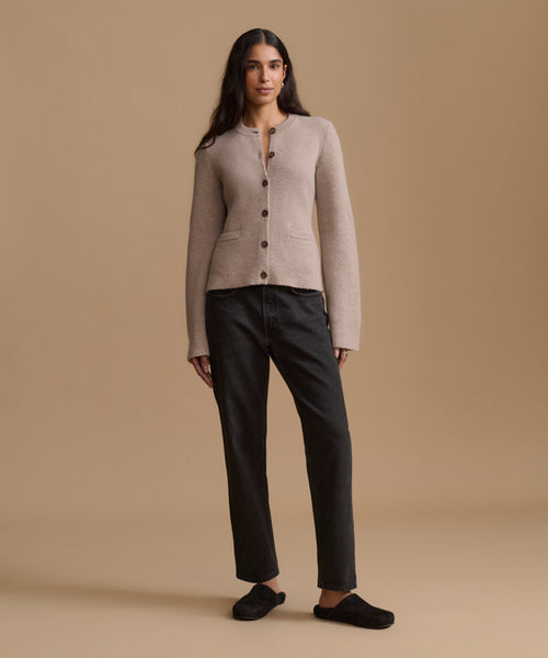 A woman stands against a warm beige studio background wearing a light taupe knit cardigan with dark buttons, black jeans, and black slip-on shoes.