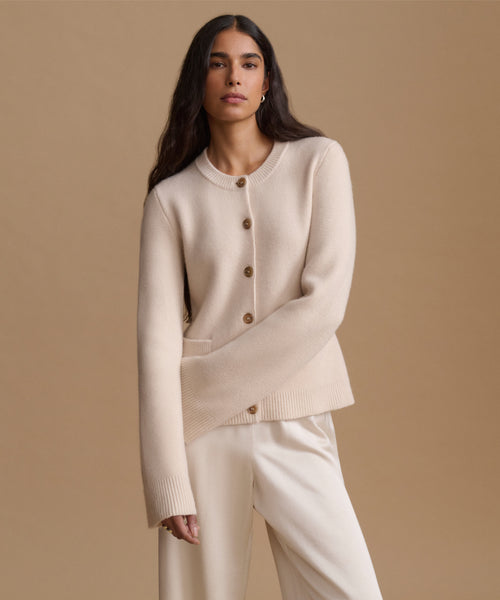 A woman modeling a cream-colored knit cardigan sweater with brown buttons, paired with matching cream trousers, against a solid beige studio background.