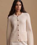 A woman wearing a cream-colored knit cardigan sweater with brown buttons, shown from the waist up against a solid beige background.
