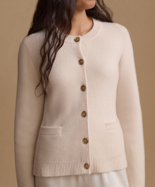 A woman wearing a cream-colored knit cardigan sweater with brown buttons, shown from the chest down against a solid beige background.