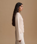 A woman in side profile wearing a cream-colored knit cardigan and matching pants against a solid beige studio background.