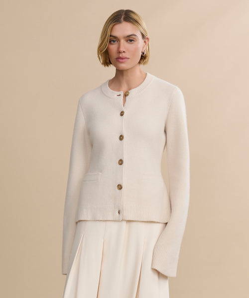 A woman with short blonde hair wears the Cooper Cardigan in Warm Ivory, featuring front pockets and a matching pleated skirt, standing against a plain beige background with a structured silhouette.
