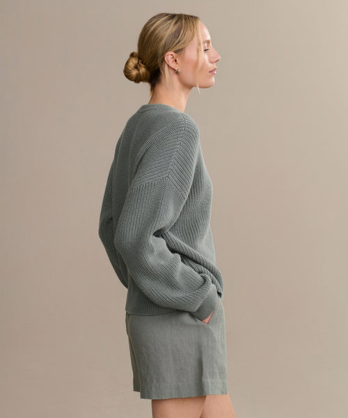 A woman with blond hair in a low bun stands in profile against a plain beige background, wearing the Cotton Hayes Crewneck Cardigan and matching gray shorts, with her hands in her pockets.