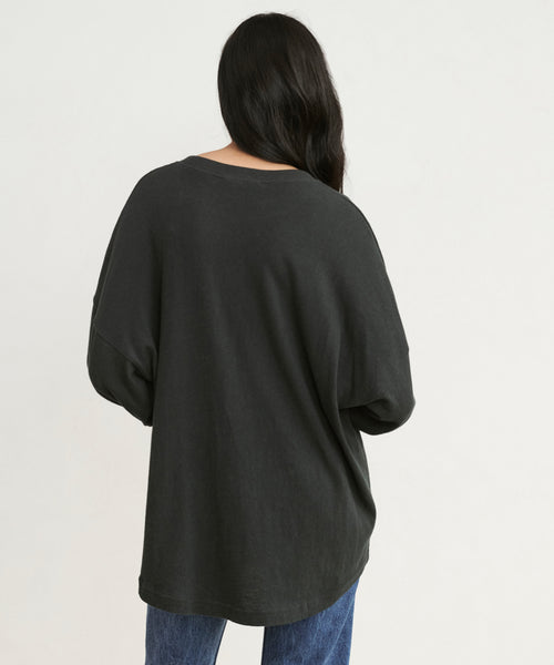A person with long dark hair stands with their back to the camera, wearing a Boyfriend Long-Sleeve Tee and blue jeans against a plain light background.