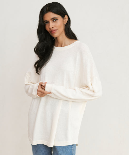 A woman with long dark hair, smiling softly and gently clasping her hands, stands against a plain background wearing the off-white Boyfriend Long-Sleeve Tee and blue jeans.