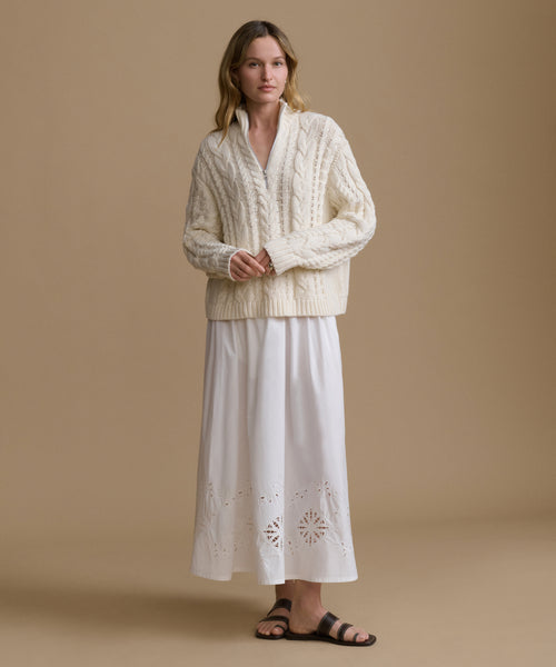 Woman wearing a cream-colored cable knit sweater over a white skirt on a beige background