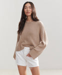 A woman with long dark hair stands against a plain light background, looking relaxed in the Cameron Crewneck sweater and white frayed-hem shorts.