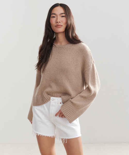 A woman with long dark hair stands against a plain light background, looking relaxed in the Cameron Crewneck sweater and white frayed-hem shorts.