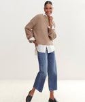 A woman smiles and poses in the Cameron Crewneck layered over a white button-up shirt, paired with blue cropped jeans and black slip-on shoes, against a plain light background.