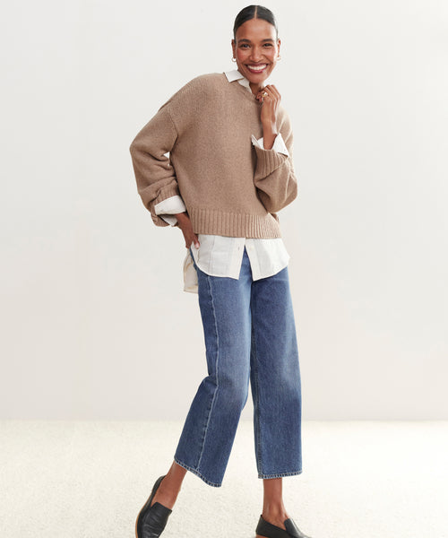 A woman smiles and poses in the Cameron Crewneck layered over a white button-up shirt, paired with blue cropped jeans and black slip-on shoes, against a plain light background.