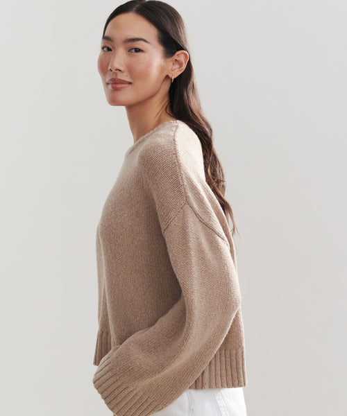 A woman with long dark hair, wearing the beige Cameron Crewneck and white pants, stands sideways and smiles softly against a plain light background.