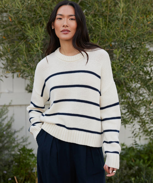 A woman with long, dark hair stands outdoors by greenery and a white fence, wearing the Chloe Crewneck—a Peruvian cotton sweater with black horizontal stripes—and dark pants, exuding a relaxed, confident vibe.