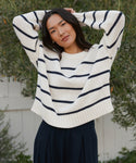 A woman with long dark hair stands outdoors by a white fence and greenery, wearing the Chloe Crewneck—a striped knit sweater crafted from soft Peruvian cotton—paired with a navy skirt. She poses with her hands resting on her head.