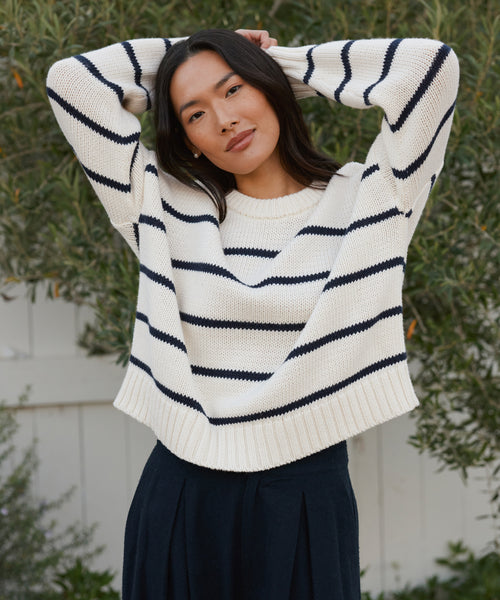 A woman with long dark hair stands outdoors by a white fence and greenery, wearing the Chloe Crewneck—a striped knit sweater crafted from soft Peruvian cotton—paired with a navy skirt. She poses with her hands resting on her head.