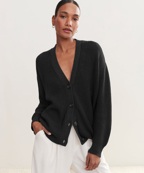 A woman stands against a plain light background, hand in pocket and relaxed, confidently wearing the Cotton Cocoon Cardigan—an organic cotton, loose-fitting black layer—paired with white pants.