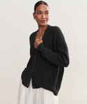 A woman models the Cotton Cocoon Cardigan in black with light pants, standing against a plain light background and looking to the side while holding the cardigan.