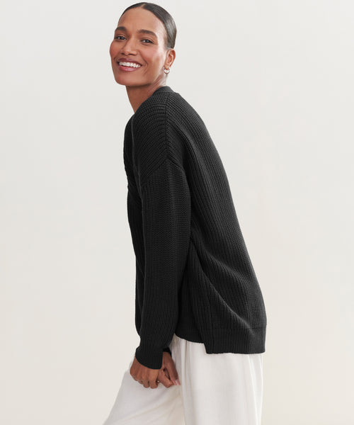 A woman in a black Cotton Cocoon Cardigan and white pants stands sideways, smiling at the camera against a plain light background. The Cotton Cocoon Cardigan is crafted from soft organic cotton for all-day comfort.