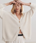 A woman in the Cotton Cocoon Cardigan—a two-button ivory cotton sweater—paired with matching pants stands with eyes closed, arms raised, and hands touching her head.