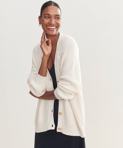 A woman smiles and looks to the side, wearing the Cotton Cocoon Cardigan in ivory, made from organic cotton knit over a dark dress, her hair pulled back and one hand gently touching her face.