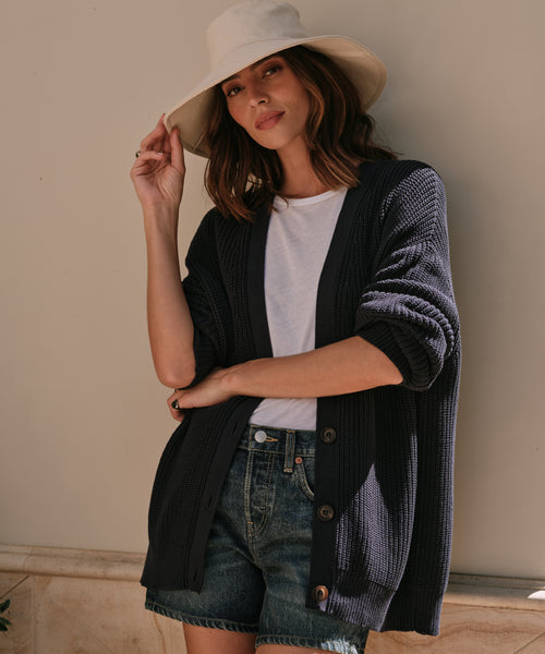 Wearing a Cotton Cocoon Cardigan, a woman in a white hat, white tee, and denim shorts stands by a light wall, smiling softly at the camera while holding her hat.