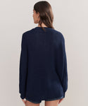 A woman with long brown hair is seen from behind wearing the Cotton Cocoon Cardigan, a loose-fitting navy blue knit sweater made from recycled polyamide, paired with denim shorts, against a plain light background.