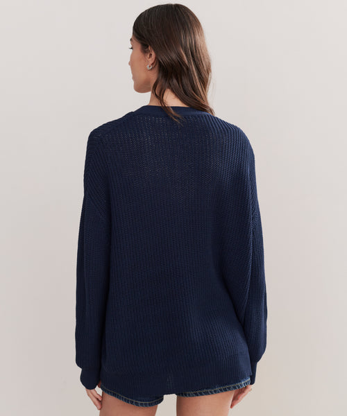A woman with long brown hair is seen from behind wearing the Cotton Cocoon Cardigan, a loose-fitting navy blue knit sweater made from recycled polyamide, paired with denim shorts, against a plain light background.