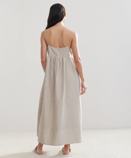 A woman with long dark hair faces away, wearing the sleeveless, ankle-length Cove Dress with thin straps and flat sandals, set against a plain light background.