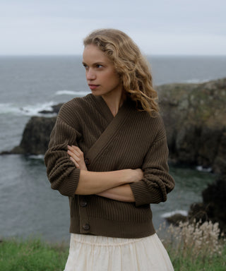 Woman wearing an olive cotton cropped cocoon cardigan standing on a rocky coastline with ocean in the background