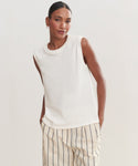 A woman with her hair pulled back stands against a plain light background, wearing the Easy Tank made of 100% cotton and cream pants with black vertical stripes. She smiles slightly, one hand in her pocket.