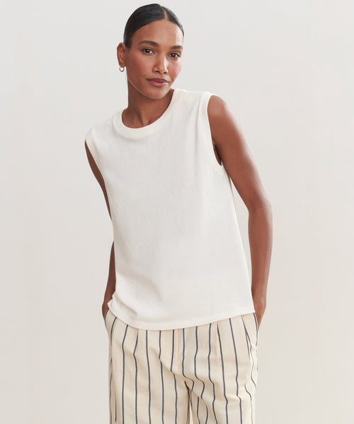 A woman with her hair pulled back stands against a plain light background, wearing the Easy Tank made of 100% cotton and cream pants with black vertical stripes. She smiles slightly, one hand in her pocket.