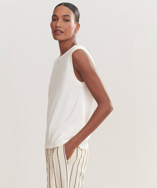 A woman, standing sideways with one hand in her pocket against a plain light background, wears the Easy Tank—a sleeveless white 100% cotton top—paired with cream pants featuring vertical black stripes.
