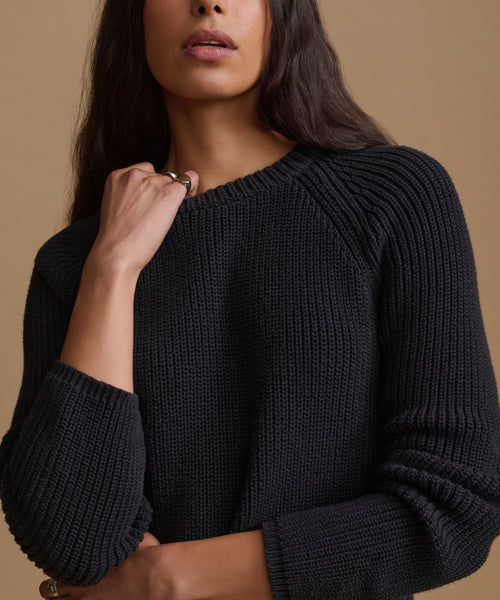 A woman modeling a black ribbed knit sweater and cream wide-leg trousers against a solid beige studio background.