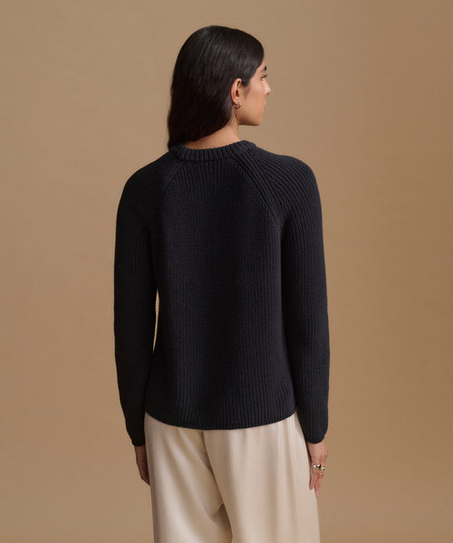 A woman shown from the back wearing a black knit sweater and light beige trousers, set against a solid warm tan studio background.