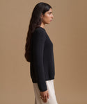 A woman in side profile wearing a black knit sweater and a cream skirt, set against a solid warm beige studio background.