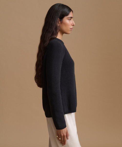 A woman in side profile wearing a black knit sweater and a cream skirt, set against a solid warm beige studio background.