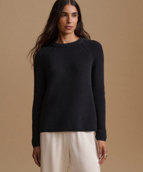 A woman wearing a black knit sweater and cream trousers, standing against a solid beige studio background.