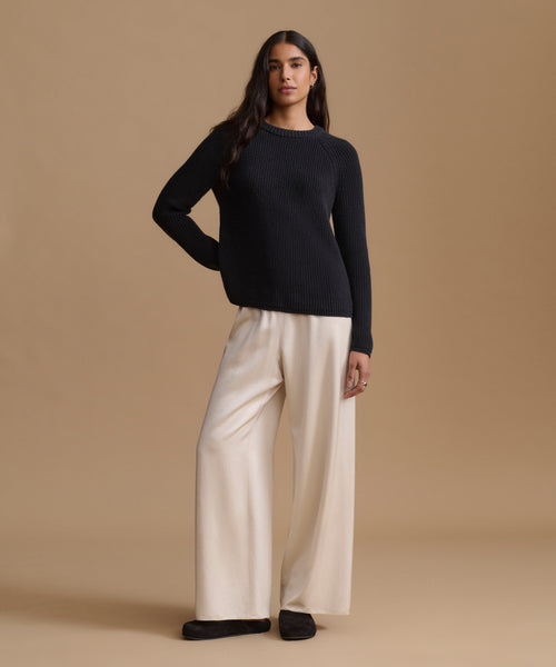 A woman modeling a black ribbed knit sweater and cream wide-leg trousers against a solid beige studio background.