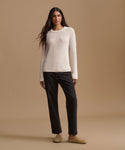 A woman stands against a warm beige studio background wearing a cream ribbed long-sleeve sweater, black straight-leg pants, and tan slip-on shoes.