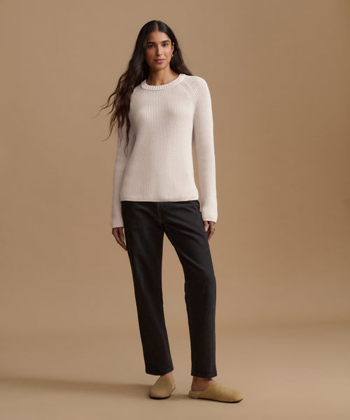 A woman stands against a warm beige studio background wearing a cream ribbed long-sleeve sweater, black straight-leg pants, and tan slip-on shoes.