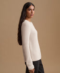 A woman shown in profile wearing a cream-colored ribbed knit sweater and dark pants against a solid beige studio background.