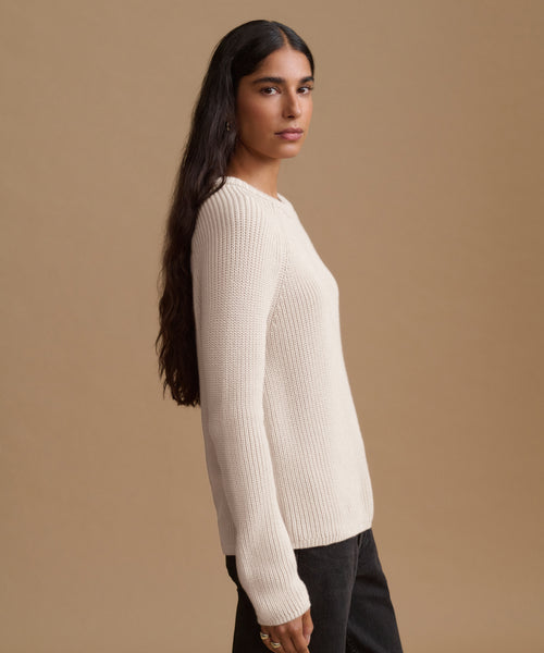 A woman shown in profile wearing a cream-colored ribbed knit sweater and dark pants against a solid beige studio background.