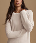 A woman wearing a cream-colored ribbed knit sweater against a solid tan background.