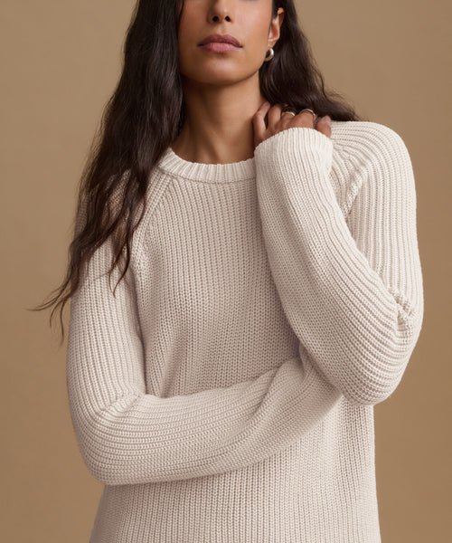A woman wearing a cream-colored ribbed knit sweater against a solid tan background.