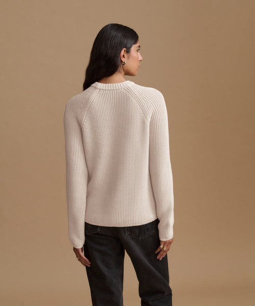 A woman shown from the back wearing a ribbed cream-colored knit sweater and dark jeans, standing against a solid tan studio background.