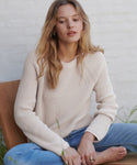 A woman with blonde hair sits barefoot on a wooden chair, wearing a cream-colored ribbed knit sweater and blue jeans against a white brick wall.