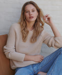 A woman wearing a beige ribbed knit sweater and blue jeans, sitting against a white textured wall.