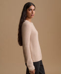 A woman shown in profile wearing a light beige ribbed knit sweater and dark pants against a solid beige studio background.