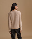 A woman shown from the back wearing a ribbed knit sweater in a light beige color, paired with dark jeans, against a solid tan studio background.